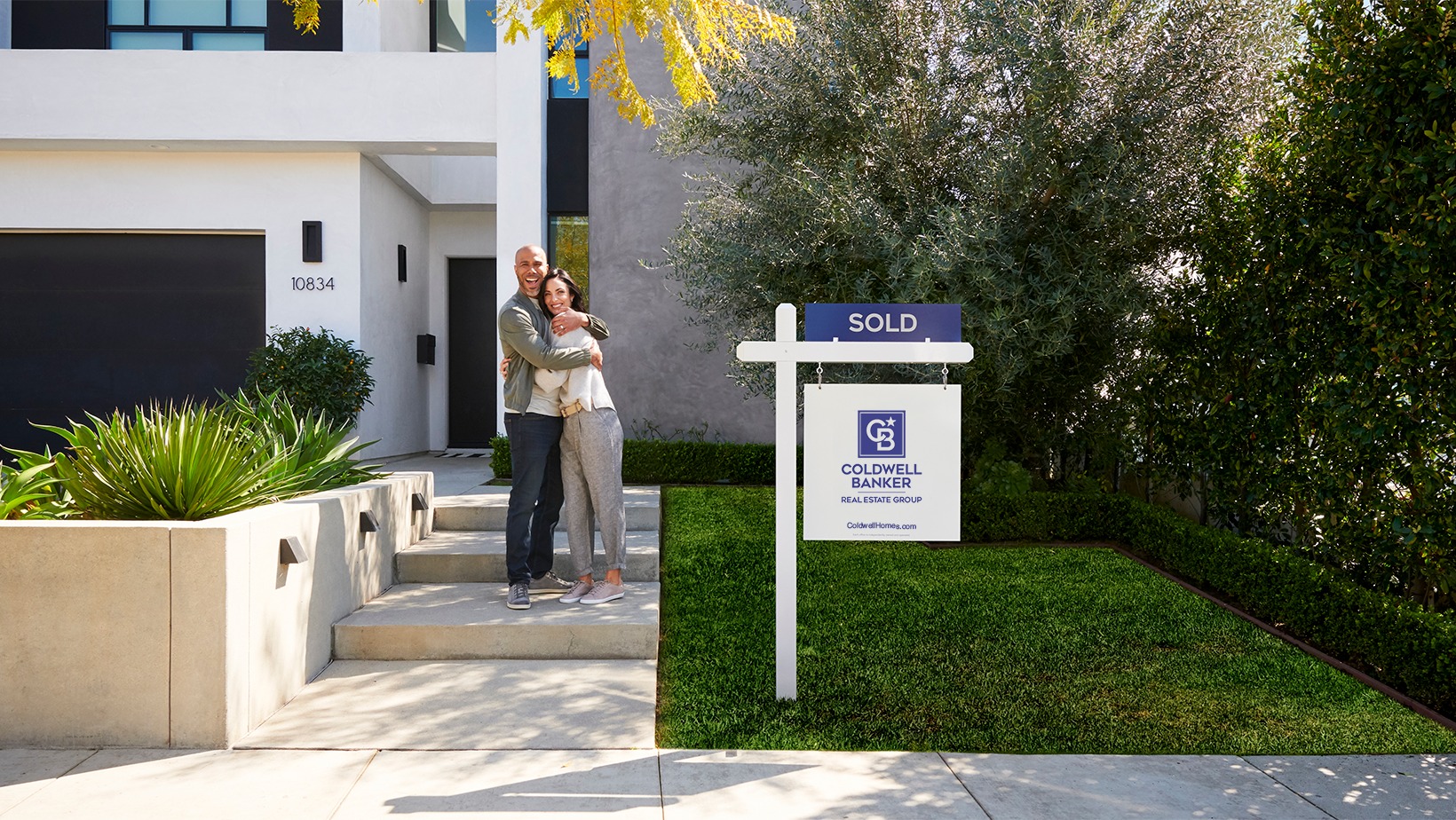 Ultimate Service satisfied clients hugging outside in front of a home with a Coldwell Banker 'sold' signage on the lawn
